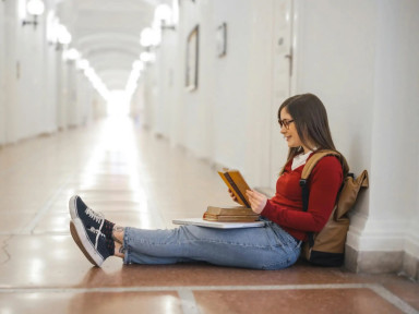 Девушка сидит в коридоре на полу с учебниками и читает